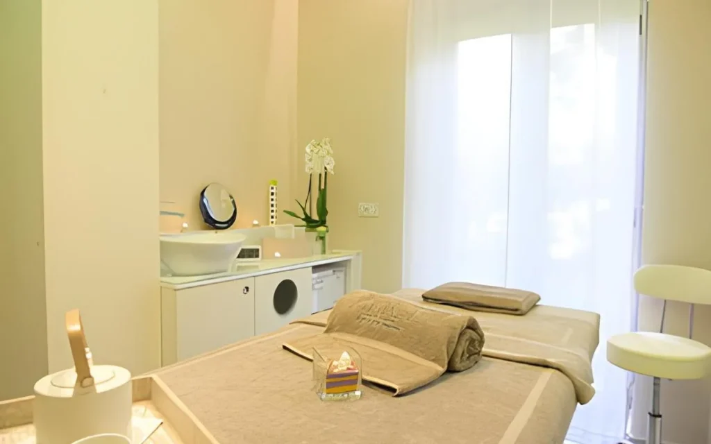 Une cabine de soin paisible chez Alice Beauty Clinique, avec une table habillée de linge taupe tout doux, des orchidées blanches sur le plan de travail, des bougies près de la vasque et des voilages qui tamisent la lumière naturelle à travers une grande fenêtre.