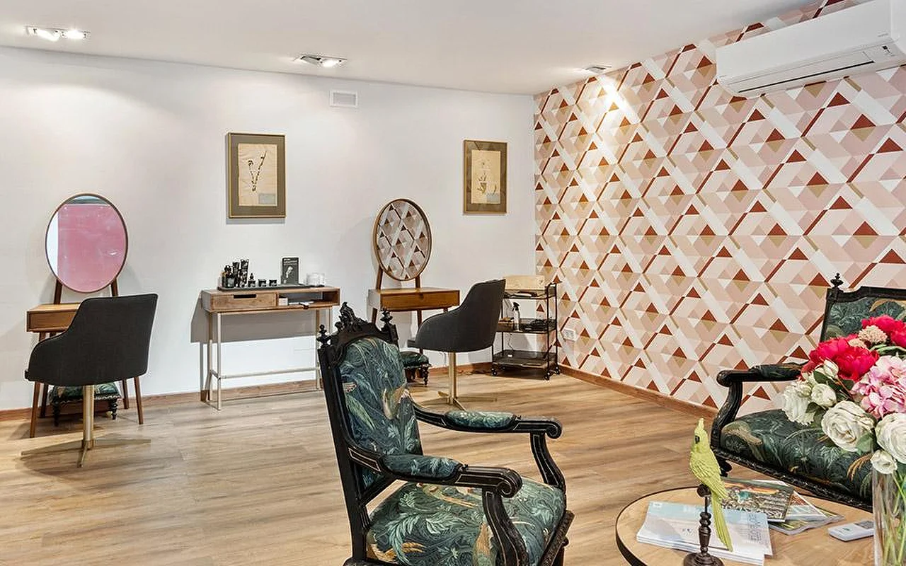 Une élégante salle de salon avec un mur d'accent géométrique rose et terracotta, deux fauteuils sculptés et travaillés tapissés d'un tissu botanique vert, des coiffeuses en bois aux miroirs ronds, et un vase de fleurs roses et rouges au premier plan.