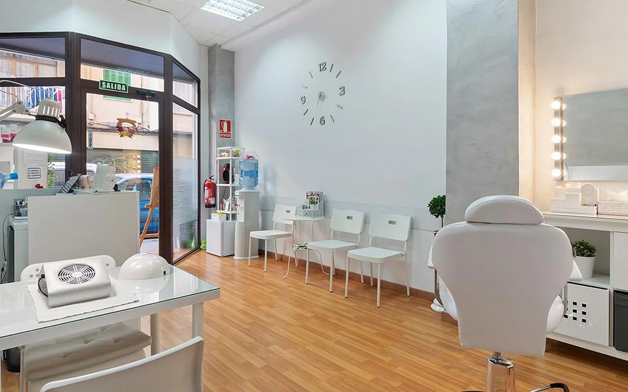 Un salon lumineux et blanc au sol en bois clair, une rangée de chaises d'attente blanches, une horloge murale minimaliste, un miroir de coiffeuse lumineux, un fauteuil de soin inclinable blanc, et un poste de manucure près de la fenêtre.