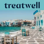 Le front de mer de la Petite Venise à Mykonos : maisons blanchies à la chaux aux balcons de bois colorés au bord de l'eau, la mer Égée turquoise au loin, et au premier plan des tables de café blanches et bleu canard installées sur une terrasse pavée.