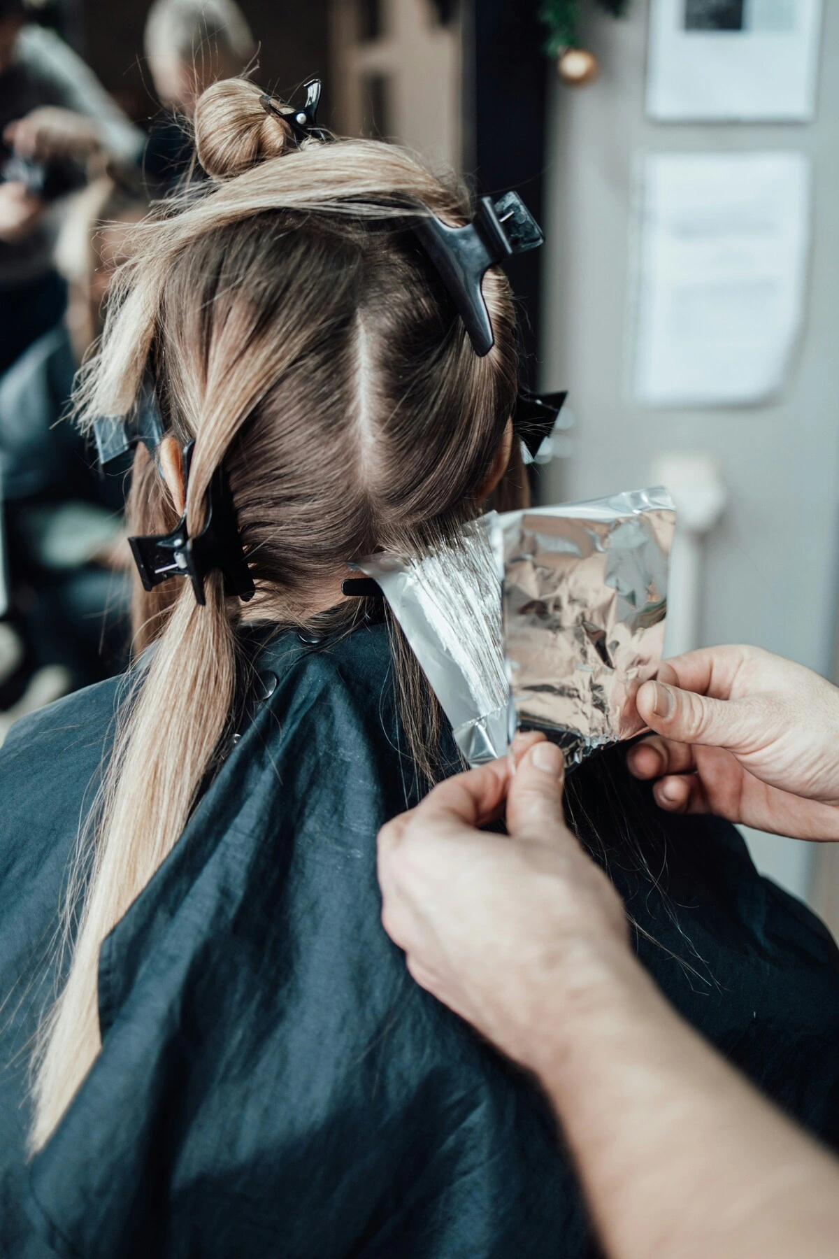 Vue de dos d'une cliente assise sur un fauteuil pendant qu'une coiffeuse place une feuille d'aluminium dans ses cheveux pour appliquer des mèches