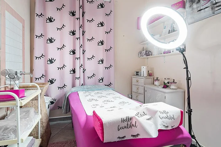 Cabine rose au Centro Estetico Karma à Olbia, avec rideaux à motifs de cils, table de soin et anneau lumineux.