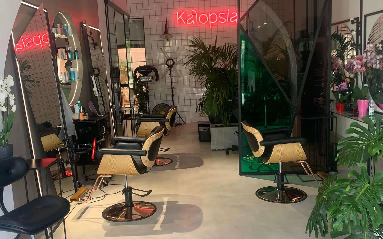 L'espace coiffage du salon Kalopsia à Ermoúpoli, Syros, avec des fauteuils en bois clair et noir, des miroirs voûtés rétroéclairés, des plantes feuillues et une enseigne néon rose indiquant Kalopsia.