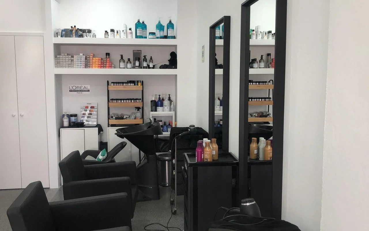 Un salon de coiffure avec des fauteuils et bacs de lavage noirs, deux hauts miroirs muraux étroits, et des étagères garnies de produits professionnels de couleur et de soin capillaire.