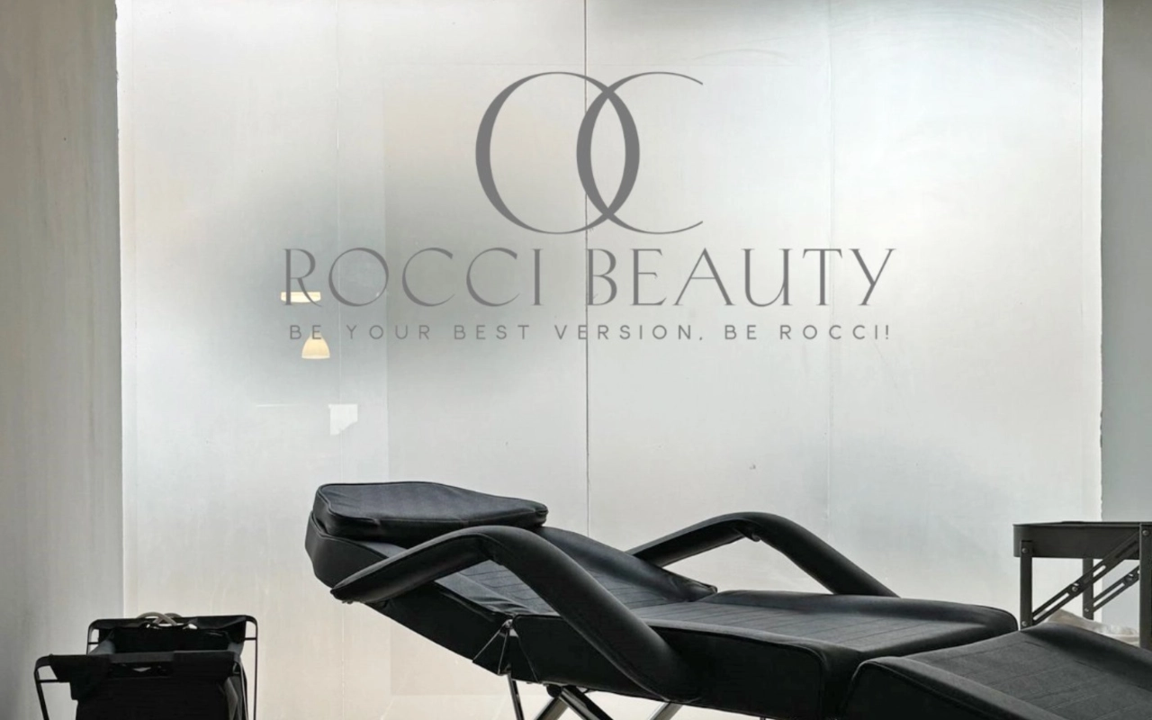 Une salle de soins avec un fauteuil inclinable noir et un petit chariot d'équipement, devant une cloison en verre dépoli portant le logo et la signature de Rocci Beauty.